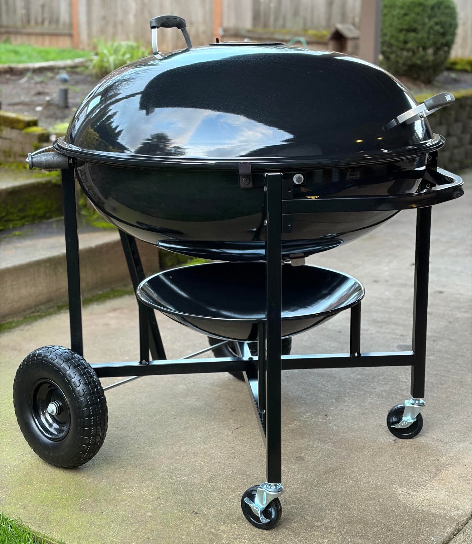 A Weber Ranch Kettle charcoal grill with a large wheels, photographed outdoors on a concrete patio with a wooden fence and grass in the background.